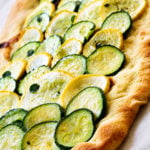 Rectangular flatbread topped with overlapping slices of green zucchini and yellow squash, baked until slightly golden, on a white surface.