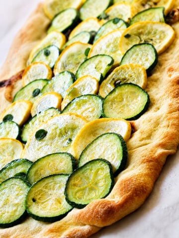 Rectangular flatbread topped with overlapping slices of green zucchini and yellow squash, baked until slightly golden, on a white surface.