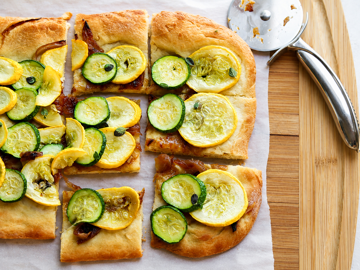 Rectangular flatbread topped with sliced yellow squash and zucchini, cut into squares. A metal pizza cutter rests nearby on a wooden board.