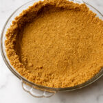 A round glass pie dish with a perfectly pressed graham cracker crust, ready for filling, sits invitingly on a white marble surface.