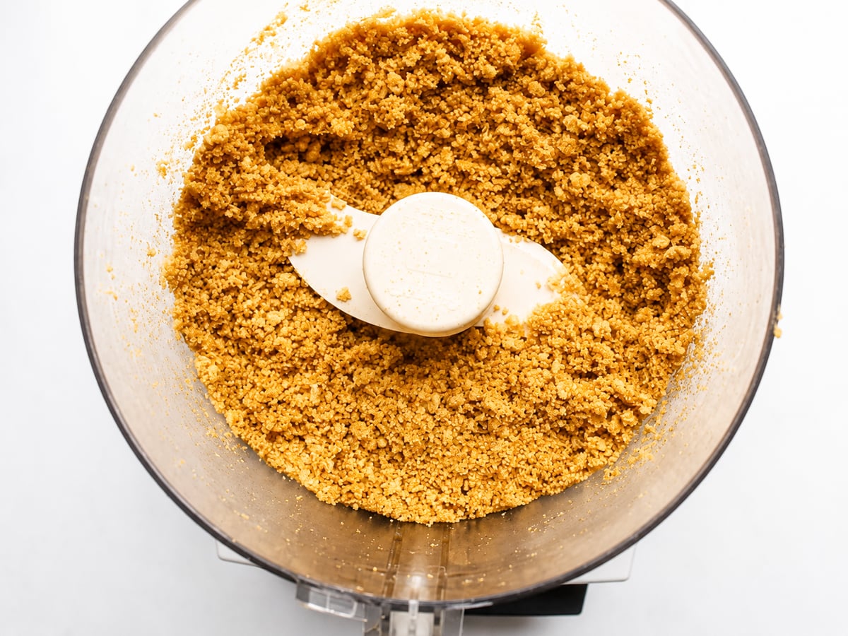 Olive Oil Graham Cracker Crust Crushed graham crackers in a food processor bowl with a white blade, forming a crumbly mixture perfect for creating a classic graham cracker crust, ready to be used in recipes like pie crust.