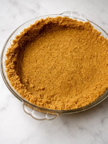 A round glass pie dish with a perfectly pressed graham cracker crust, ready for filling, sits invitingly on a white marble surface.