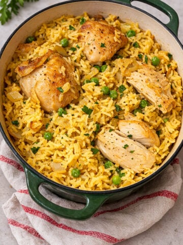 A green Dutch oven filled with yellow rice, green peas, and browned chicken pieces, garnished with chopped parsley, sits on a beige surface with a red-striped cloth-this is a Very Easy One Pot Chicken and Rice meal.