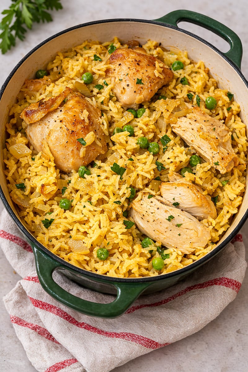 A green Dutch oven filled with yellow rice, green peas, and browned chicken pieces, garnished with chopped parsley, sits on a beige surface with a red-striped cloth&mdash;this is a Very Easy One Pot Chicken and Rice meal.