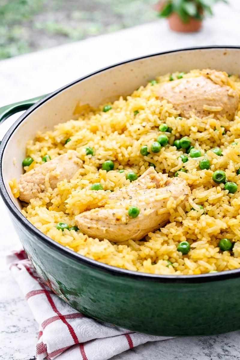 A green Dutch oven filled with yellow rice, green peas, and pieces of cooked chicken breast sits on a striped kitchen towel&mdash;a Very Easy One Pot Chicken and Rice dish that's both colorful and inviting.