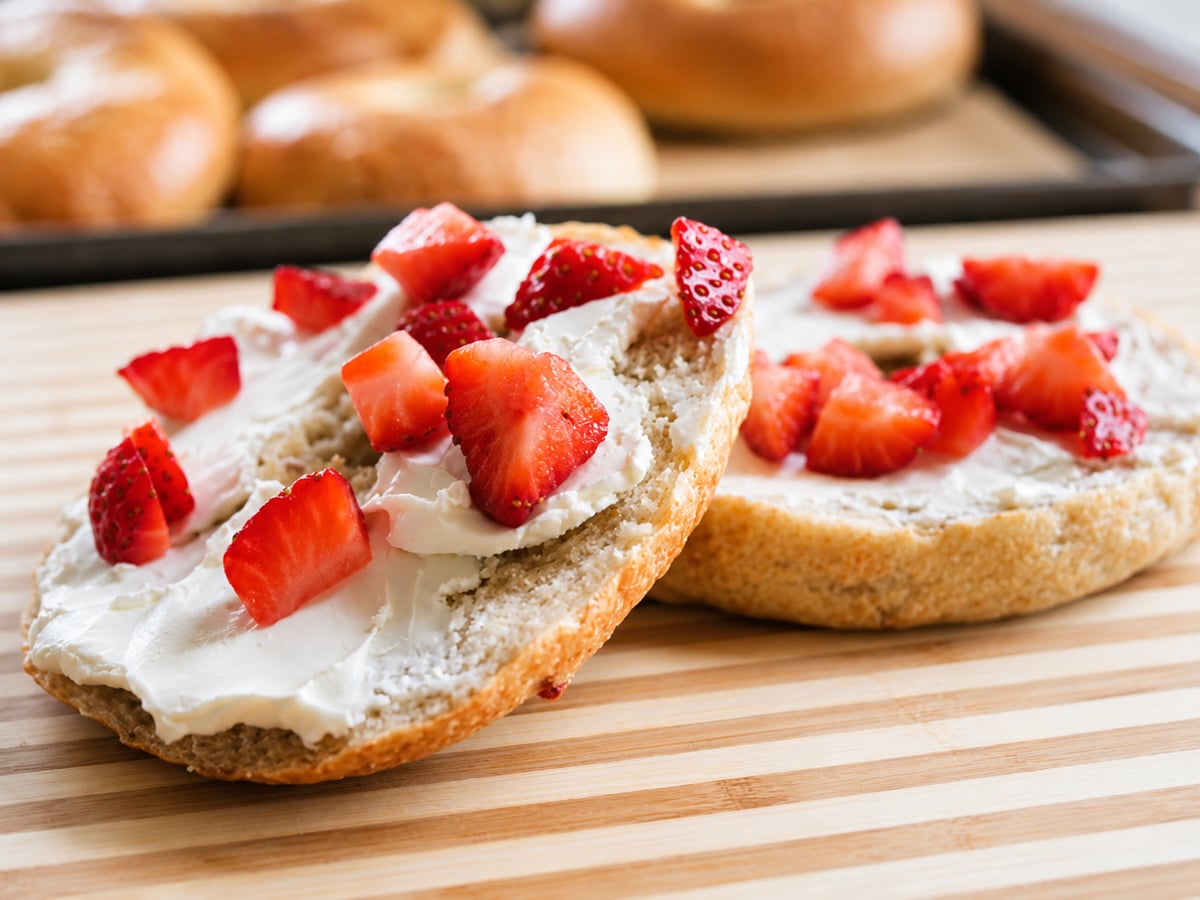 A sliced Strawberry Bagel with cream cheese and fresh strawberry pieces sits on a wooden striped surface, with whole bagels blurred in the background.