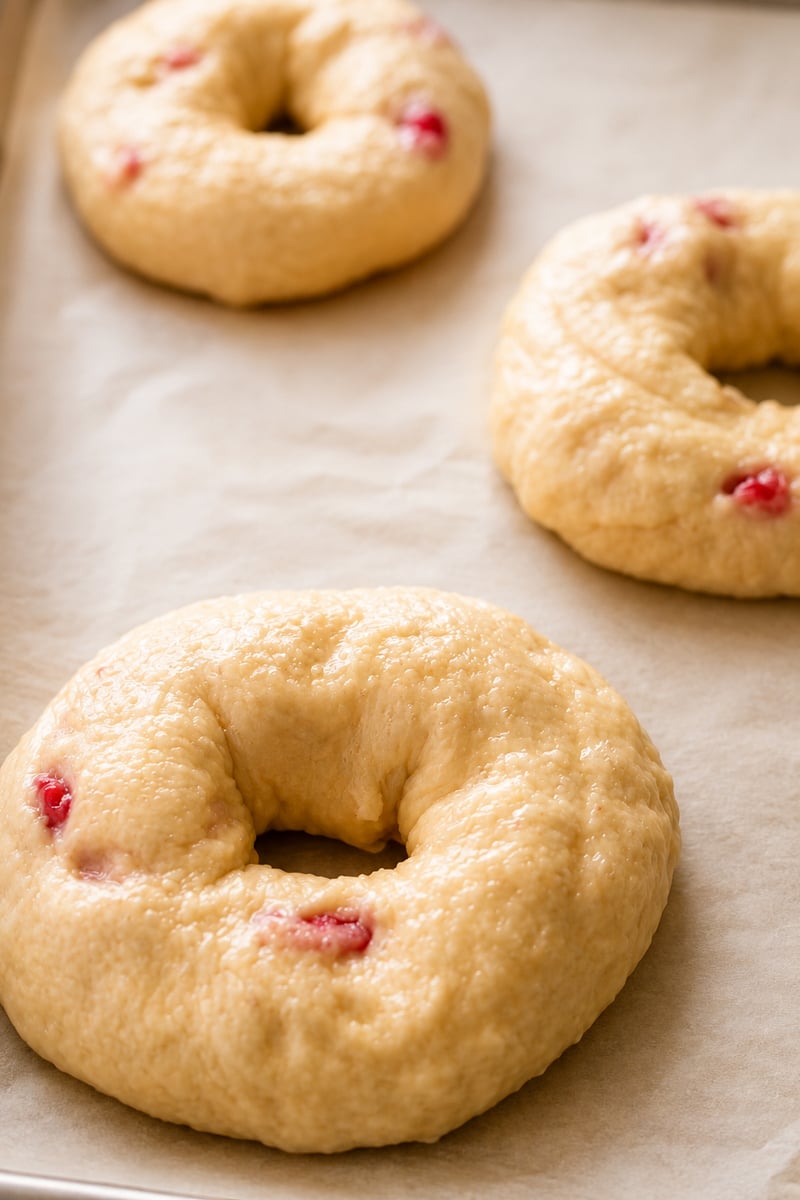 Unbaked strawberry bagels, dotted with a few red fruit pieces, sit on a parchment-lined baking sheet, ready to be baked.