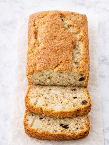 A loaf of Banana Pear Bread with Dark Chocolate Chips boasts a golden-brown crust, partially sliced to reveal a moist interior with chunks of fruit and chocolate, resting on parchment paper atop a light surface.