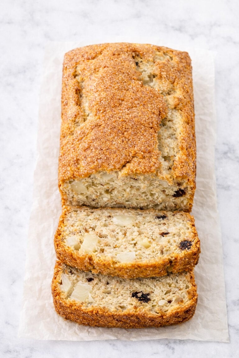 A loaf of Banana Pear Bread with Dark Chocolate Chips boasts a golden-brown crust, partially sliced to reveal a moist interior with chunks of fruit and chocolate, resting on parchment paper atop a light surface.