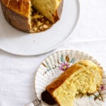 A slice of Sicilian Orange Sweet Bread panettone on a decorative plate with a vintage knife, next to the remaining loaf on a white plate, all set atop a crisp white tablecloth.