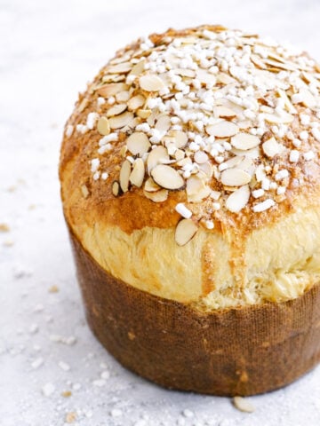 A tall, round loaf of Sicilian Orange Sweet Bread, golden-brown and topped with sliced almonds and pearl sugar, sits in a brown paper liner on a light, textured surface.