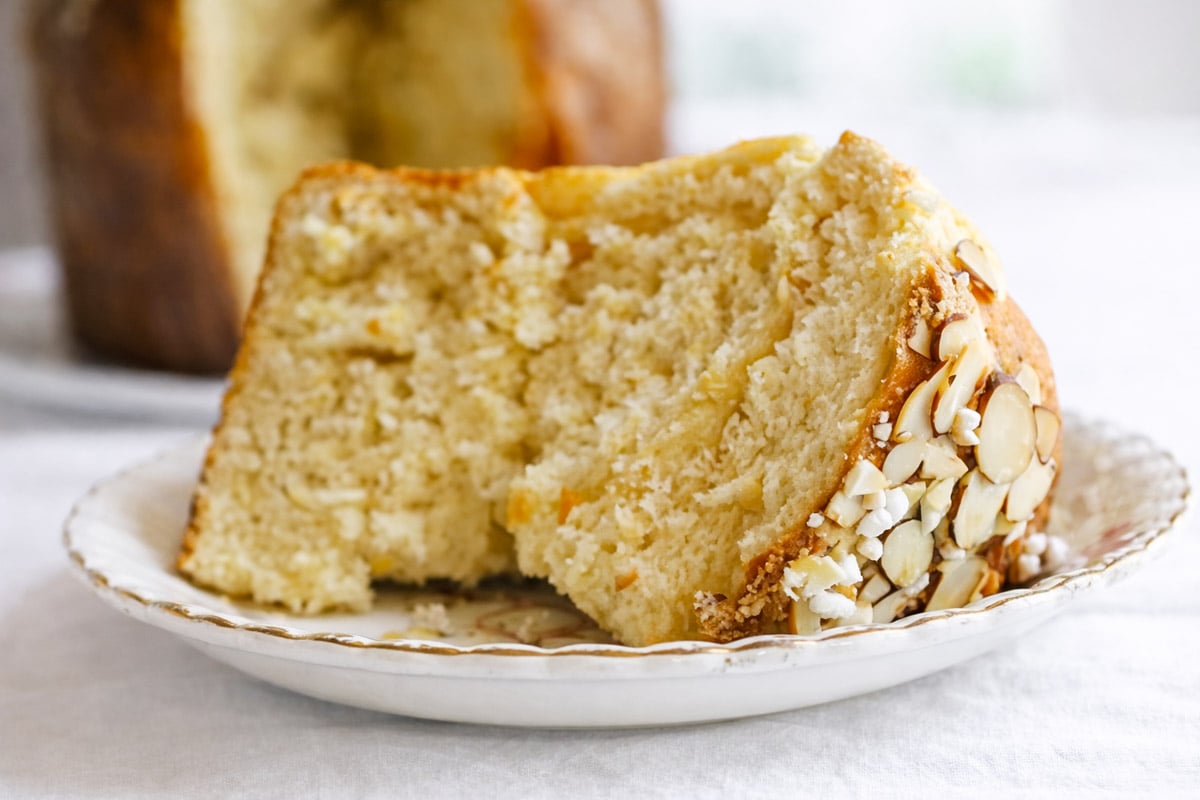 A close-up of a slice of sweet, fluffy Sicilian Orange Sweet Bread garnished with sliced almonds on the crust, served on a decorative plate with a blurred background.