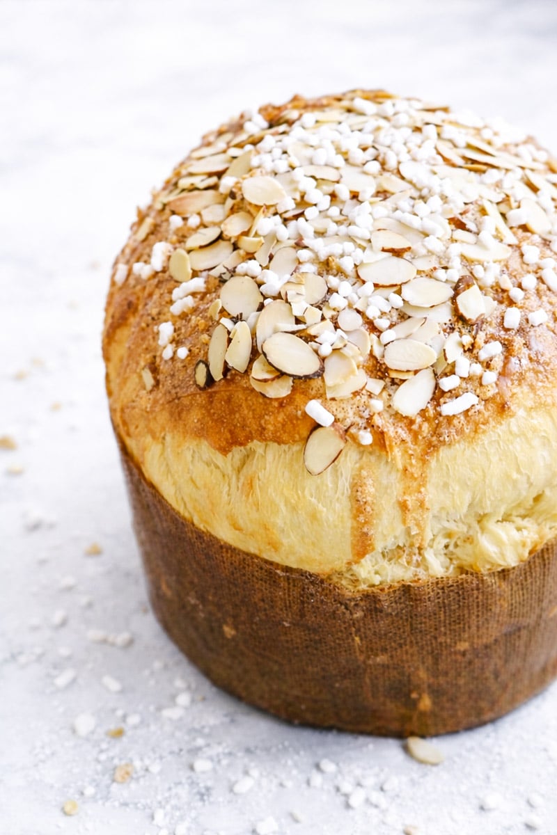 A tall, round loaf of Sicilian Orange Sweet Bread, golden-brown and topped with sliced almonds and pearl sugar, sits in a brown paper liner on a light, textured surface.