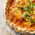 Close-up of a baked quiche in a glass pie dish, showing a golden, flaky crust with a filling of melted cheese, leafy greens, and savory bits of ham and goat cheese.
