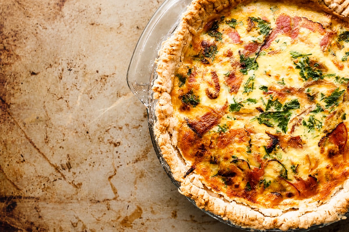 A close-up of a ham and goat cheese tart in a glass pie dish, showing a golden, flaky crust and creamy filling with pieces of Vidalia onion and fresh herbs, set on a rustic, lightly stained baking surface.