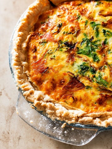 Close-up of a baked quiche in a glass pie dish, showing a golden, flaky crust with a filling of melted cheese, leafy greens, and savory bits of ham and goat cheese.