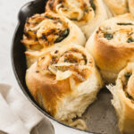 Golden, fluffy Caramelized Vidalia Onion Rolls filled with savory onions and herbs, baked in a white cast iron skillet. A soft, airy texture is visible where one roll has been removed. A beige cloth is nearby.