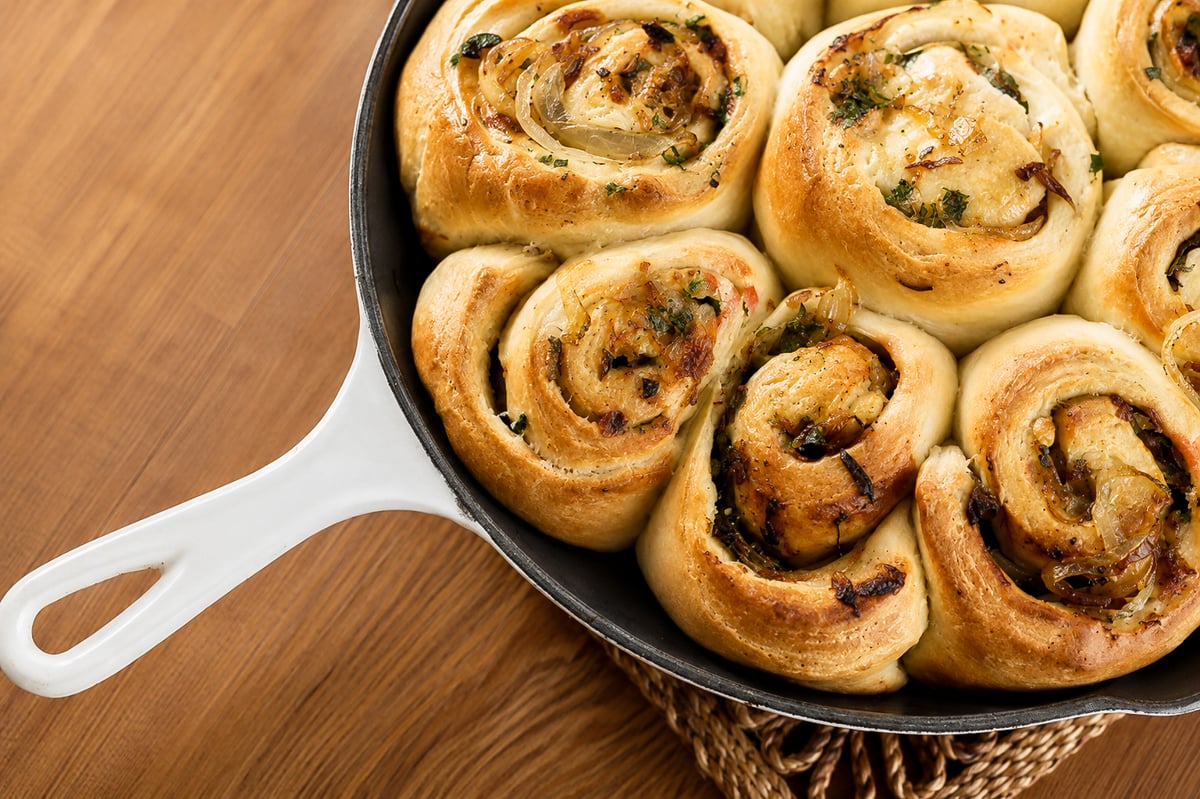 Caramelized Vidalia Onion Rolls A close-up of golden, freshly baked Caramelized Vidalia Onion Rolls with herbs in a white-handled skillet on a wooden surface.