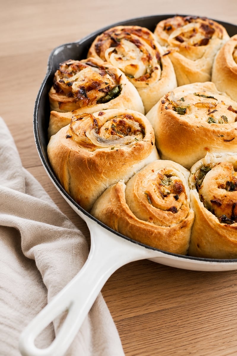 Caramelized Vidalia Onion Rolls A white skillet filled with golden-brown Caramelized Vidalia Onion Rolls, swirled with herbs and melted cheese, sits on a light wooden table next to a beige cloth napkin.