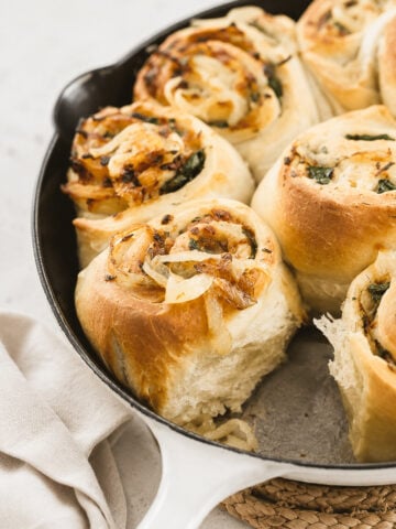Golden, fluffy Caramelized Vidalia Onion Rolls filled with savory onions and herbs, baked in a white cast iron skillet. A soft, airy texture is visible where one roll has been removed. A beige cloth is nearby.