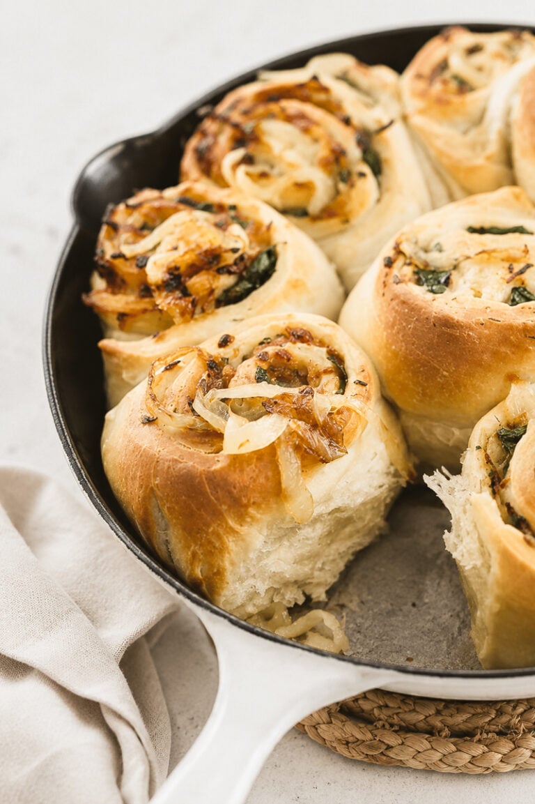 Golden, fluffy Caramelized Vidalia Onion Rolls filled with savory onions and herbs, baked in a white cast iron skillet. A soft, airy texture is visible where one roll has been removed. A beige cloth is nearby.