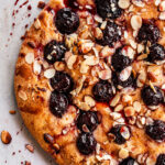 A close-up of a Cherry Almond Focaccia tart topped with dark cherries and sliced almonds, resting on a light-colored surface with crumbs and juice splatters.