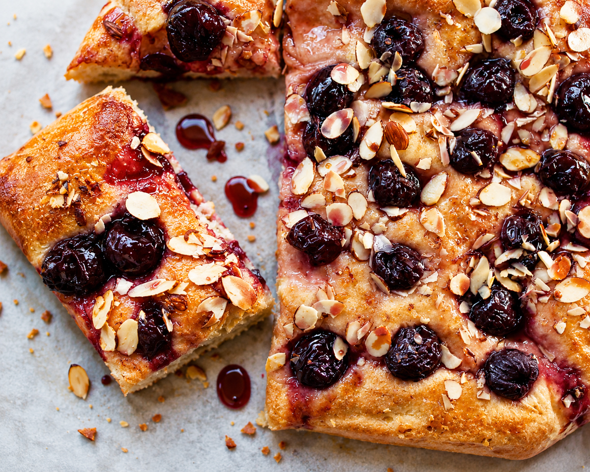 A rectangular Cherry Almond Focaccia with a golden crust, topped with sliced almonds and whole cherries. One slice is cut and slightly pulled away, revealing the soft, airy texture inside. Syrup and crumbs dot the surface.