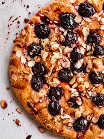A close-up of a Cherry Almond Focaccia tart topped with dark cherries and sliced almonds, resting on a light-colored surface with crumbs and juice splatters.
