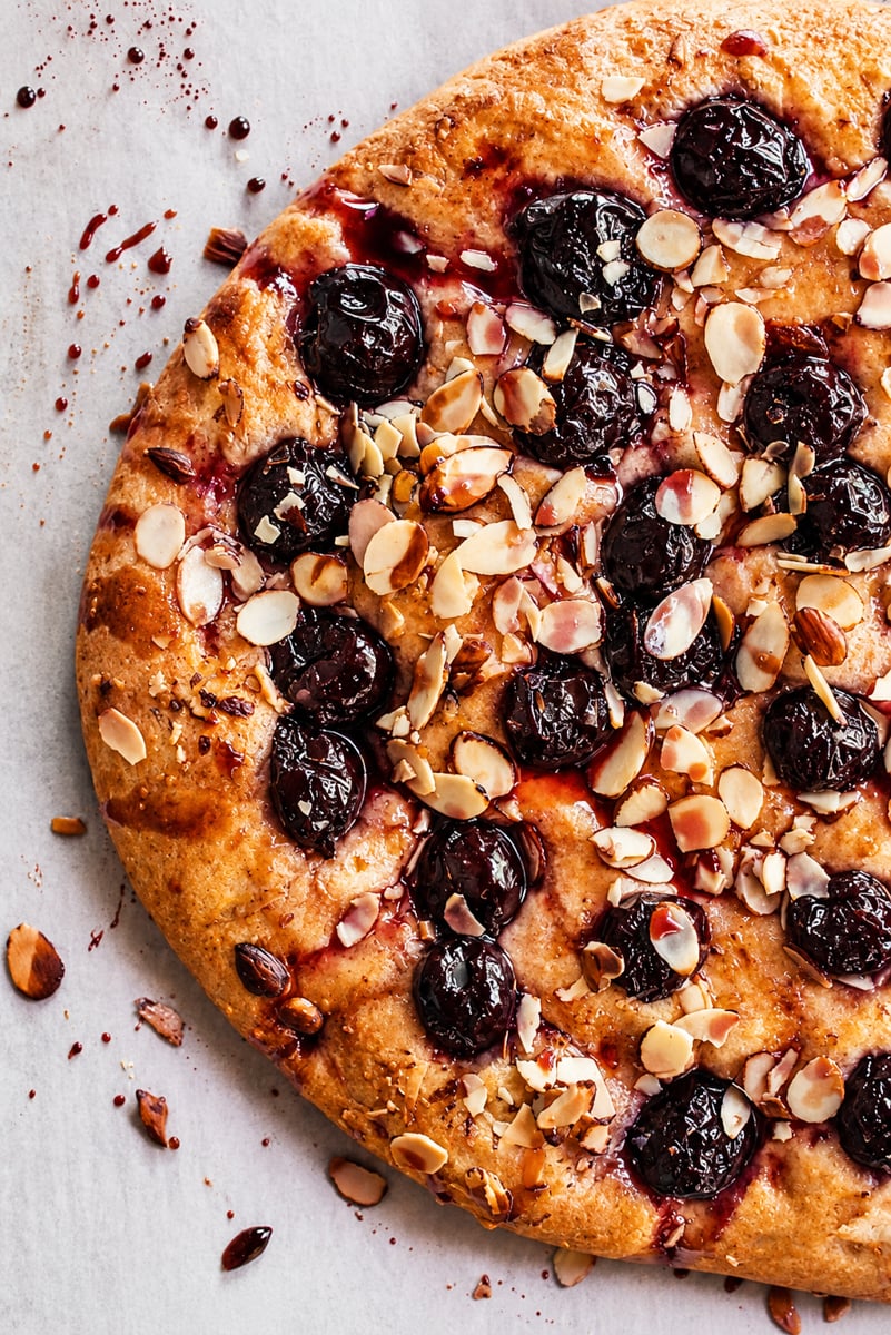 A close-up of a Cherry Almond Focaccia tart topped with dark cherries and sliced almonds, resting on a light-colored surface with crumbs and juice splatters.