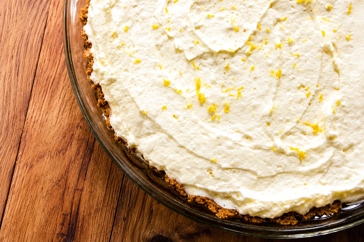 A creamy No-Bake Limoncello Cheesecake with a crumbly crust in a glass dish, topped with grated lemon zest, sits on a wooden surface.