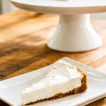 A slice of creamy No-Bake Limoncello Cheesecake with a graham cracker crust is served on a white square plate, with the rest of the cheesecake on a cake stand in the background.