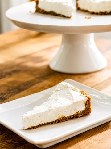 A slice of creamy No-Bake Limoncello Cheesecake with a graham cracker crust is served on a white square plate, with the rest of the cheesecake on a cake stand in the background.