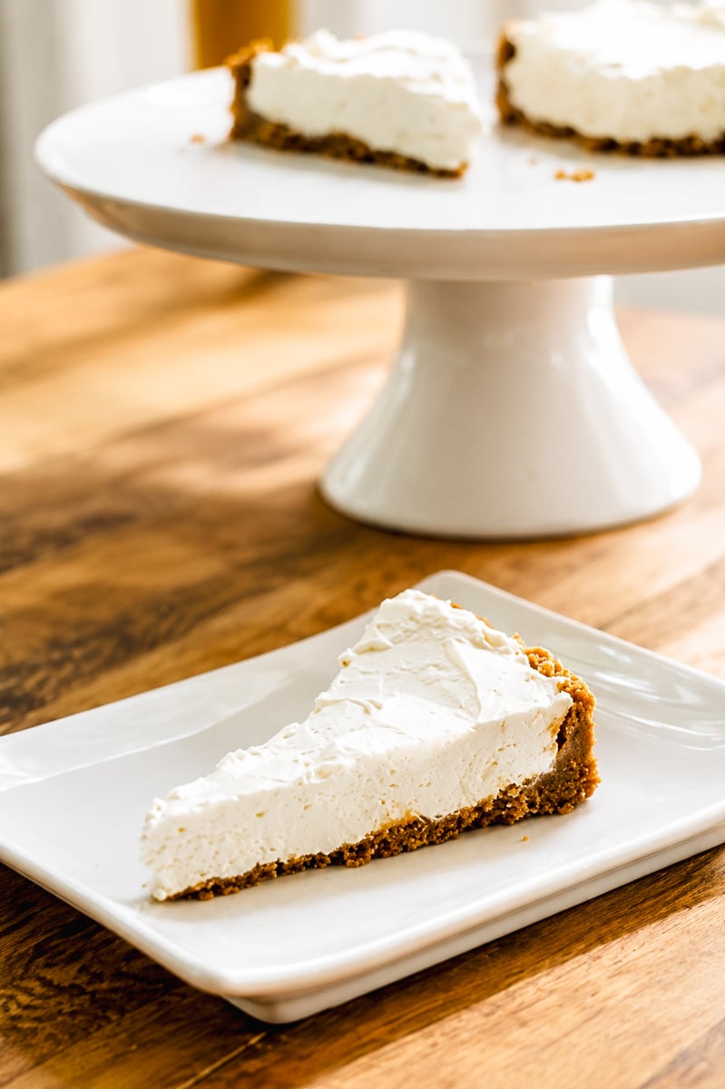 A slice of creamy No-Bake Limoncello Cheesecake with a graham cracker crust is served on a white square plate, with the rest of the cheesecake on a cake stand in the background.