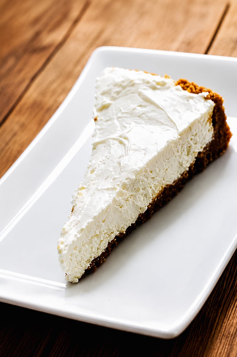 A slice of creamy No-Bake Limoncello Cheesecake with a graham cracker crust sits on a white rectangular plate atop a wooden surface.