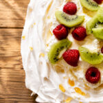 A close-up of a classic pavlova dessert topped with sliced kiwi and whole raspberries on swirled whipped cream, drizzled with honey, all set on a rustic wooden surface.