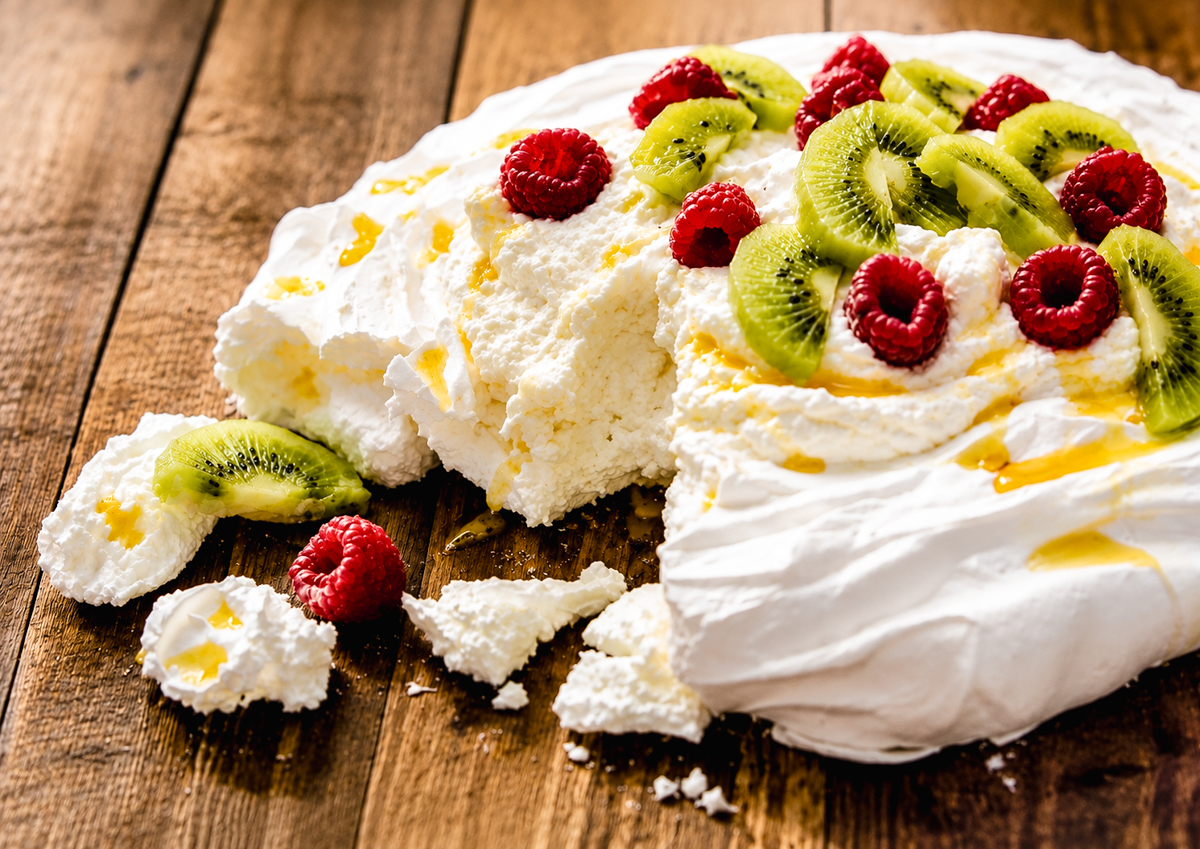 A classic Pavlova topped with sliced kiwifruit and raspberries, drizzled with honey, sits on a rustic wooden table with some broken pavlova pieces scattered around the edges.