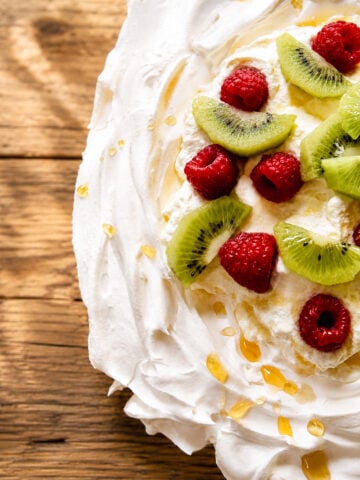 A close-up of a classic pavlova dessert topped with sliced kiwi and whole raspberries on swirled whipped cream, drizzled with honey, all set on a rustic wooden surface.