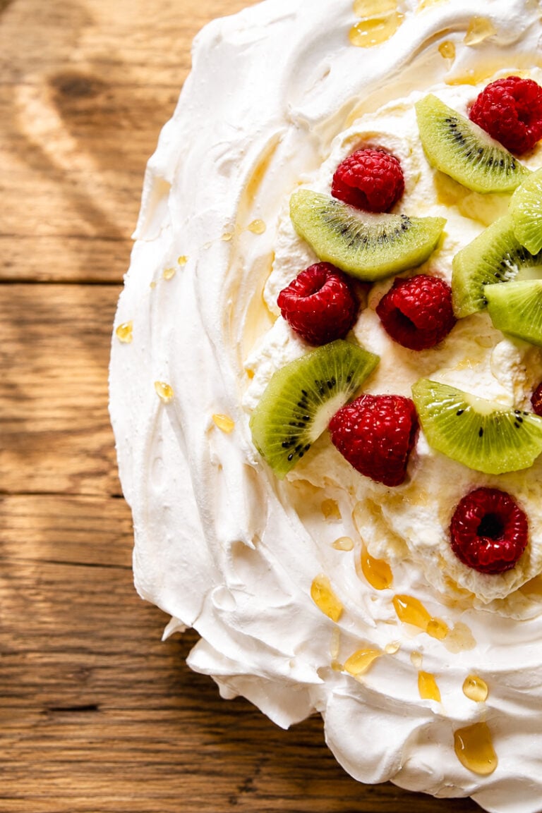 A close-up of a classic pavlova dessert topped with sliced kiwi and whole raspberries on swirled whipped cream, drizzled with honey, all set on a rustic wooden surface.