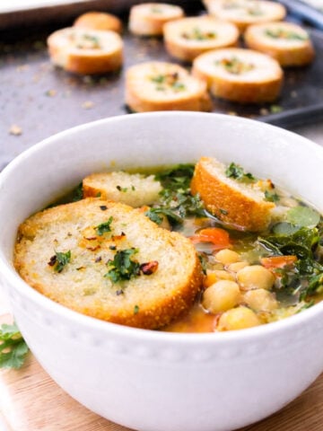 A white bowl of kale and chickpea soup is topped with toasted bread slices and garnished with herbs. In the background, more toasted bread slices rest on a baking sheet.