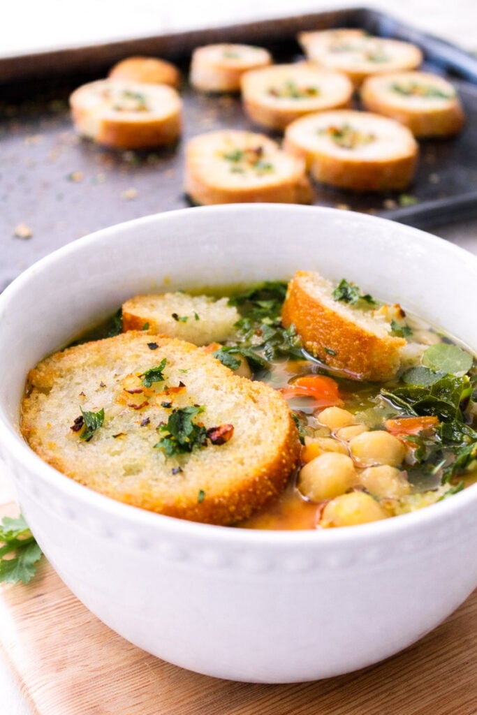 A white bowl of kale and chickpea soup is topped with toasted bread slices and garnished with herbs. In the background, more toasted bread slices rest on a baking sheet.