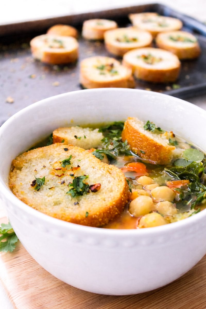 A white bowl of kale and chickpea soup is topped with toasted bread slices and garnished with herbs. In the background, more toasted bread slices rest on a baking sheet.