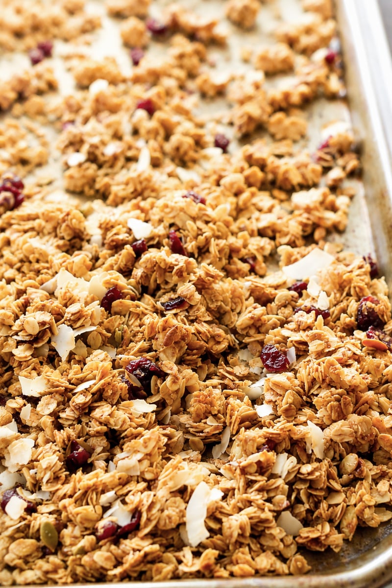 A close-up of super easy cranberry coconut granola on a baking sheet, featuring oats, dried cranberries, coconut flakes, and seeds, golden brown and freshly baked.