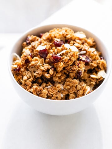 A white bowl filled with super easy cranberry coconut granola, featuring oats, dried cranberries, almond slices, and a touch of coconut, sits on a white surface.