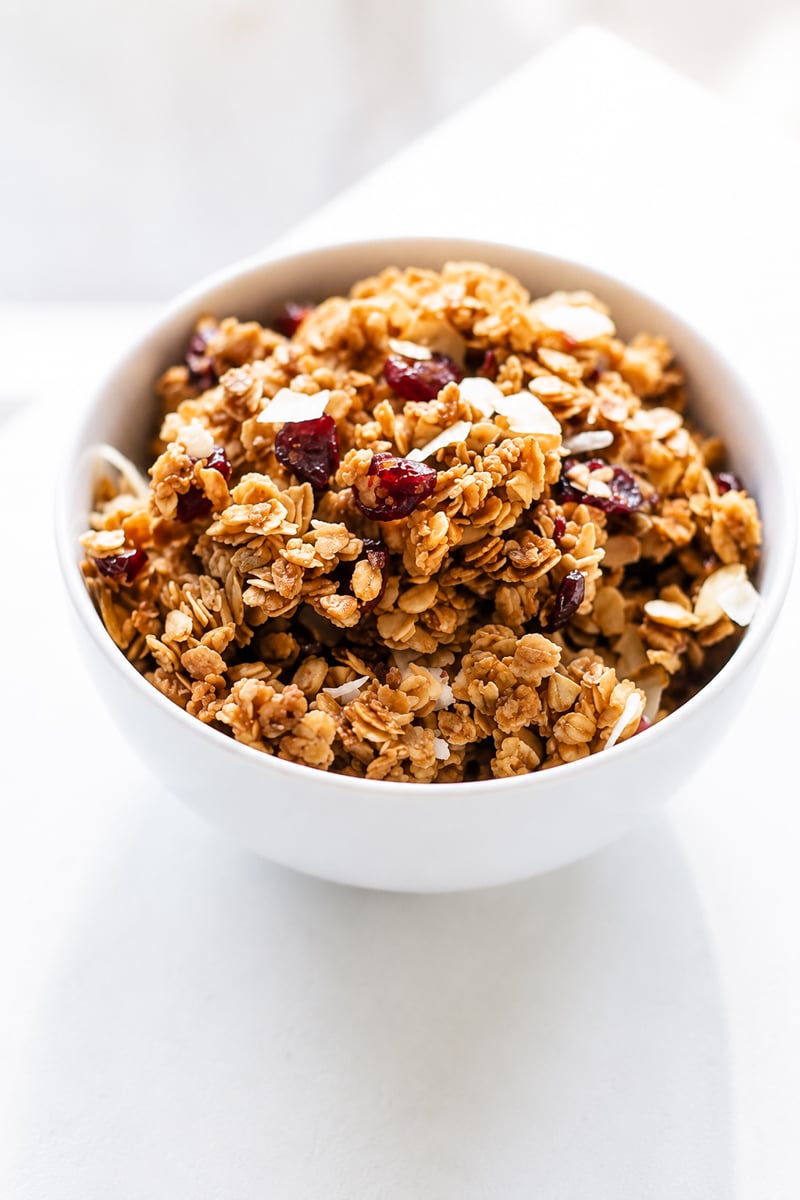 A white bowl filled with super easy cranberry coconut granola, featuring oats, dried cranberries, almond slices, and a touch of coconut, sits on a white surface.