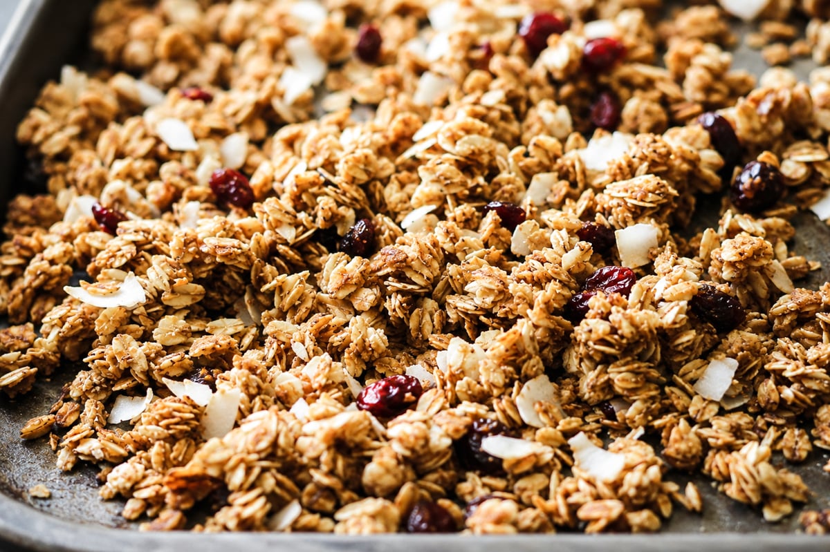 A close-up of super easy cranberry coconut granola with oats, coconut flakes, and dried cranberries spread on a baking sheet.
