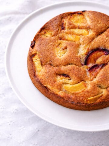 A round, golden-brown cake with visible slices of peaches and plums on top, displayed on a white plate set on a light, textured tablecloth.