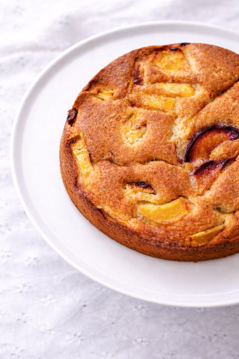 A round, golden-brown cake with visible slices of peaches and plums on top, displayed on a white plate set on a light, textured tablecloth.