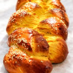 A golden-brown braided pumpkin challah bread topped with sesame seeds on parchment paper.