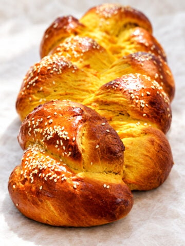 A golden-brown braided pumpkin challah bread topped with sesame seeds on parchment paper.