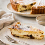 A slice of ricotta and raisin cake, reminiscent of a classic ricotta pie, is topped with powdered sugar and sits on a ceramic plate with a fork. In the background, the rest of the cake waits, partially sliced next to a beige napkin.